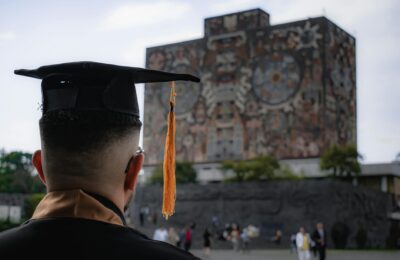 Students walking through a modern Middle Eastern university campus with palm-lined plaza, contemporary library building, and graduates in regalia
