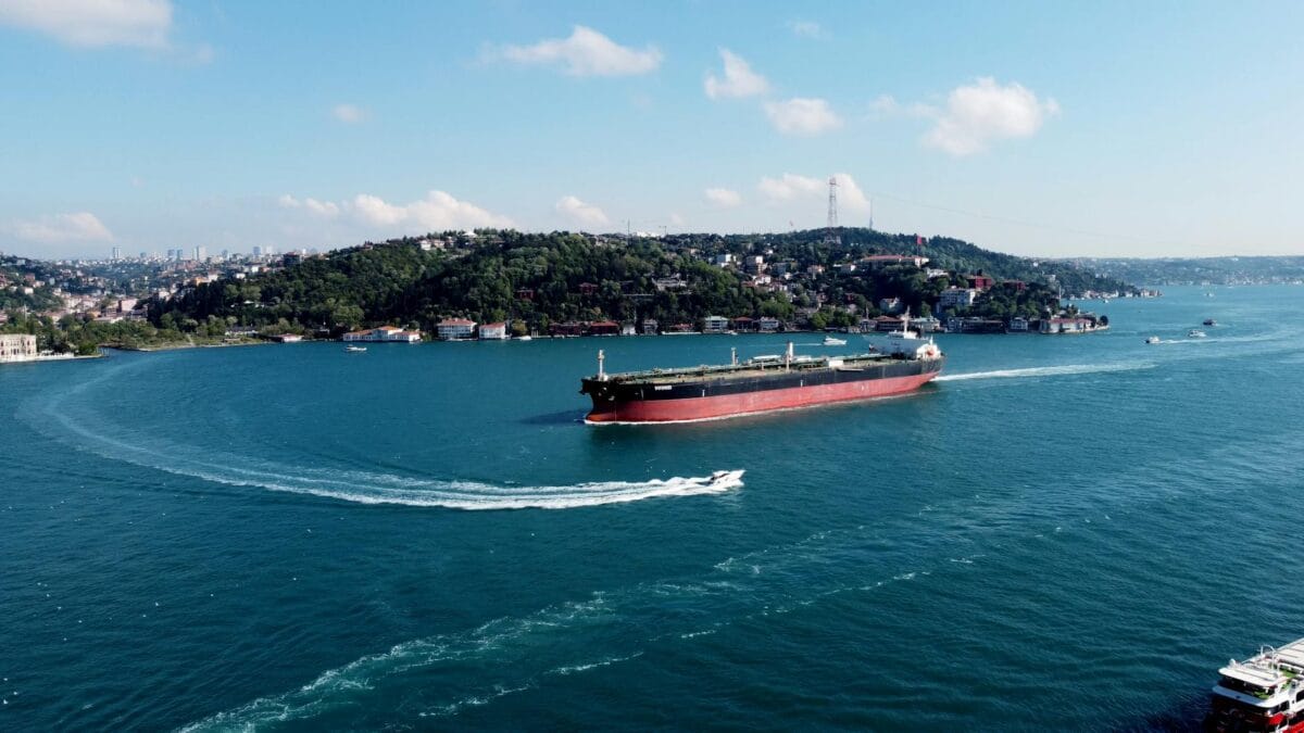 An aerial view of a stranded oil tanker idling at the entrance of a narrow shipping channel near the Strait of Hormuz, with naval escort vessels visible in the distance